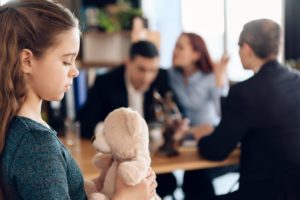 Girl holding her teddy bear while her parents are arguing at the background.