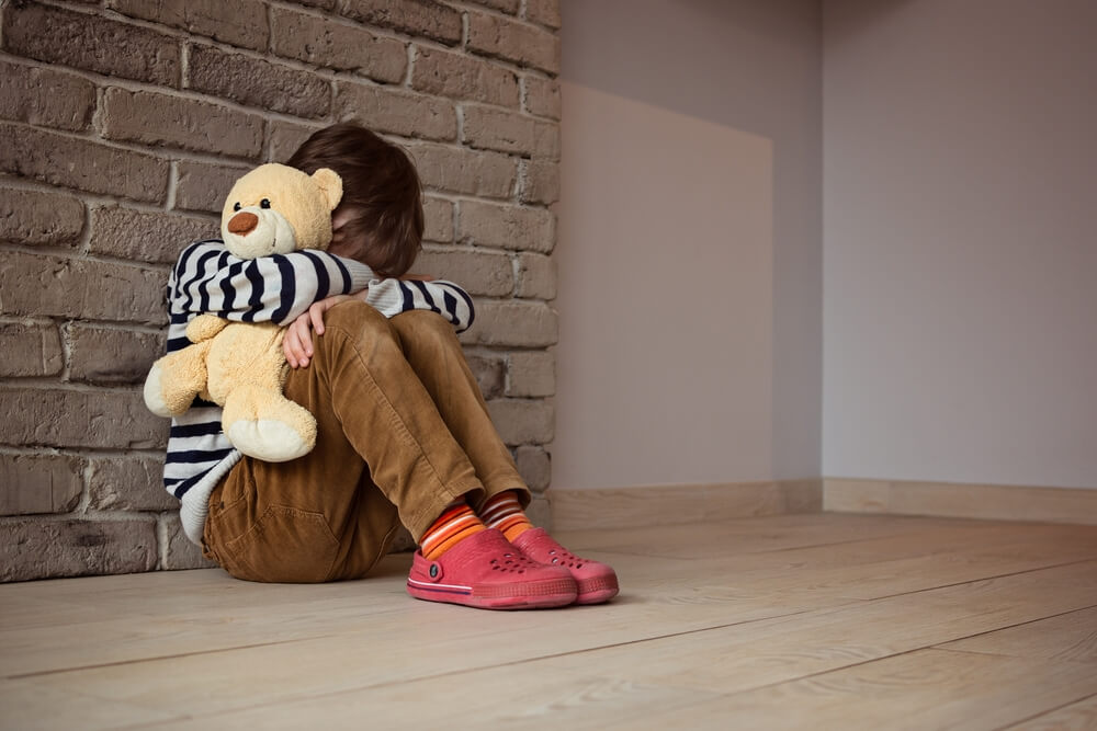 Sad boy sitting against the wall with his teddy bear.