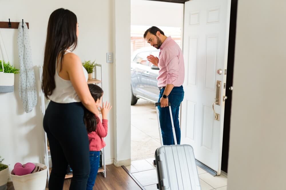 Sad father using a suitcase waving goodbye to her little daughter and mother while leaving the house after divorce.